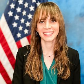 A blonde woman wearing a green shirt and black blazer smiles in front of an American flag.