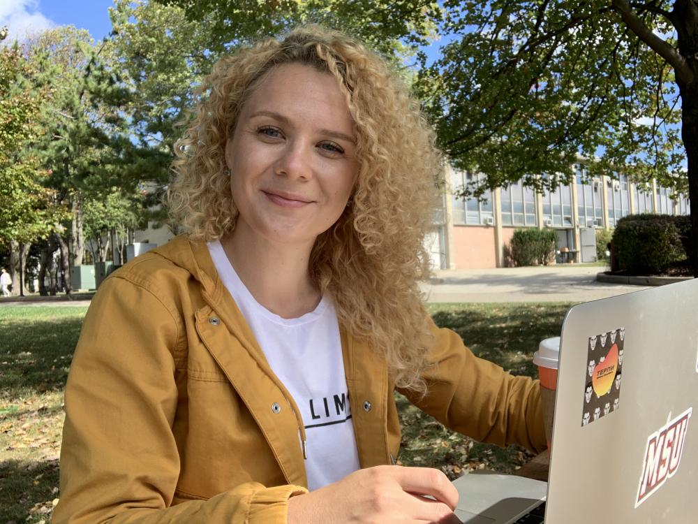 A white woman with curly blonde hair sits and smiles in a grassy outdoor area with a laptop.