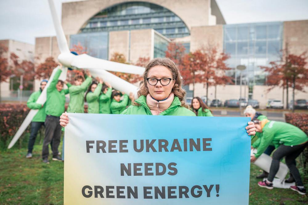 A white woman with blonde hair stands in front of a group of people wearing matching green jackets and supporting a wind turbine; she holds a sign reading "Free Ukraine needs green energy now!"