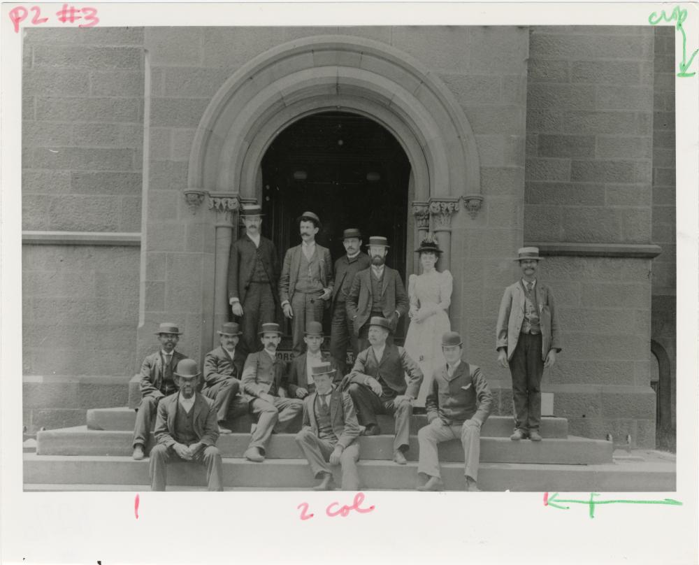 A black and white photo og a group of people in Victorian clothes standing in front of a building.